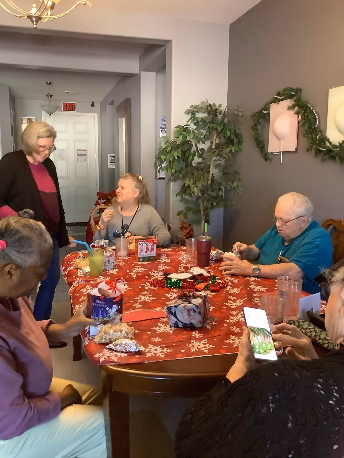 Seniors and a staff member gathered around a decorated table sharing snacks and gifts in a communal dining room.