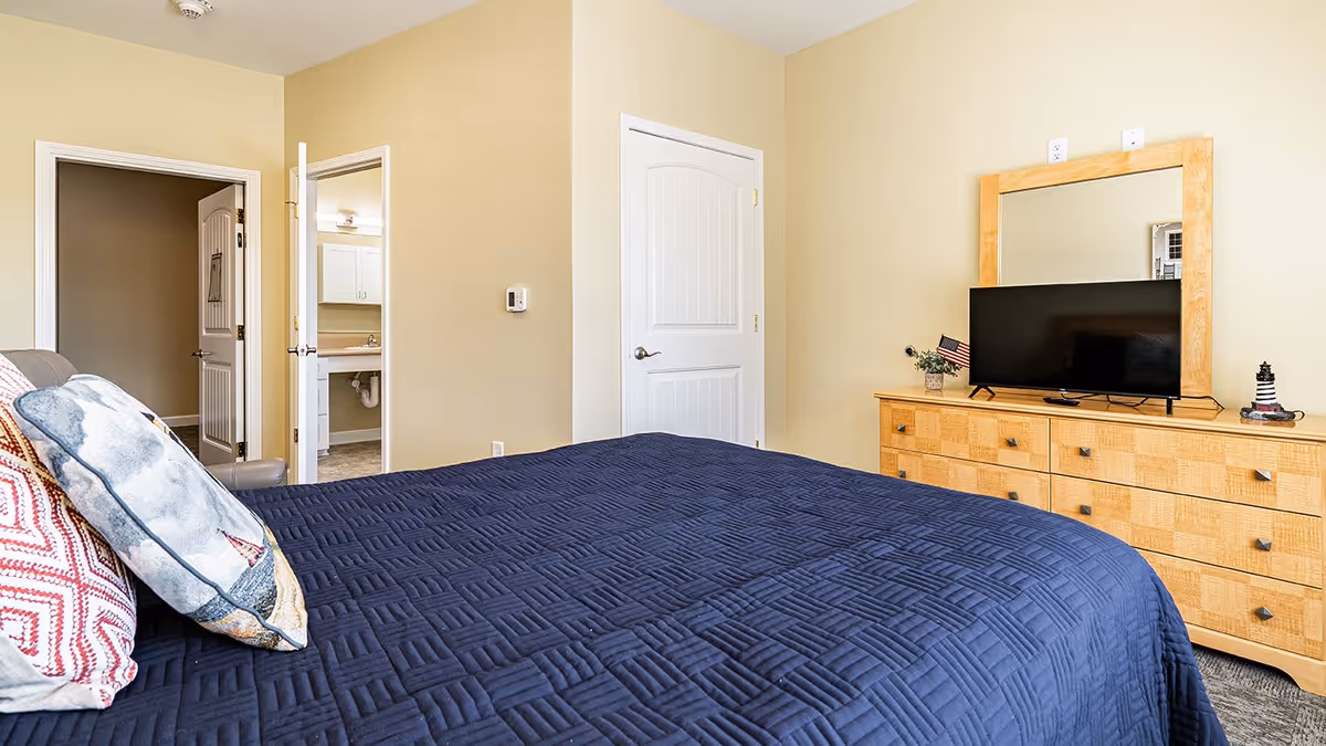 A bedroom with a bed covered in a dark blue quilt and decorative pillows. There is a wooden dresser with a large mirror and a flat-screen TV on top. Two doors are visible, one leading to a bathroom with a sink and mirror, and the other likely a closet or another room. The walls are painted light beige.