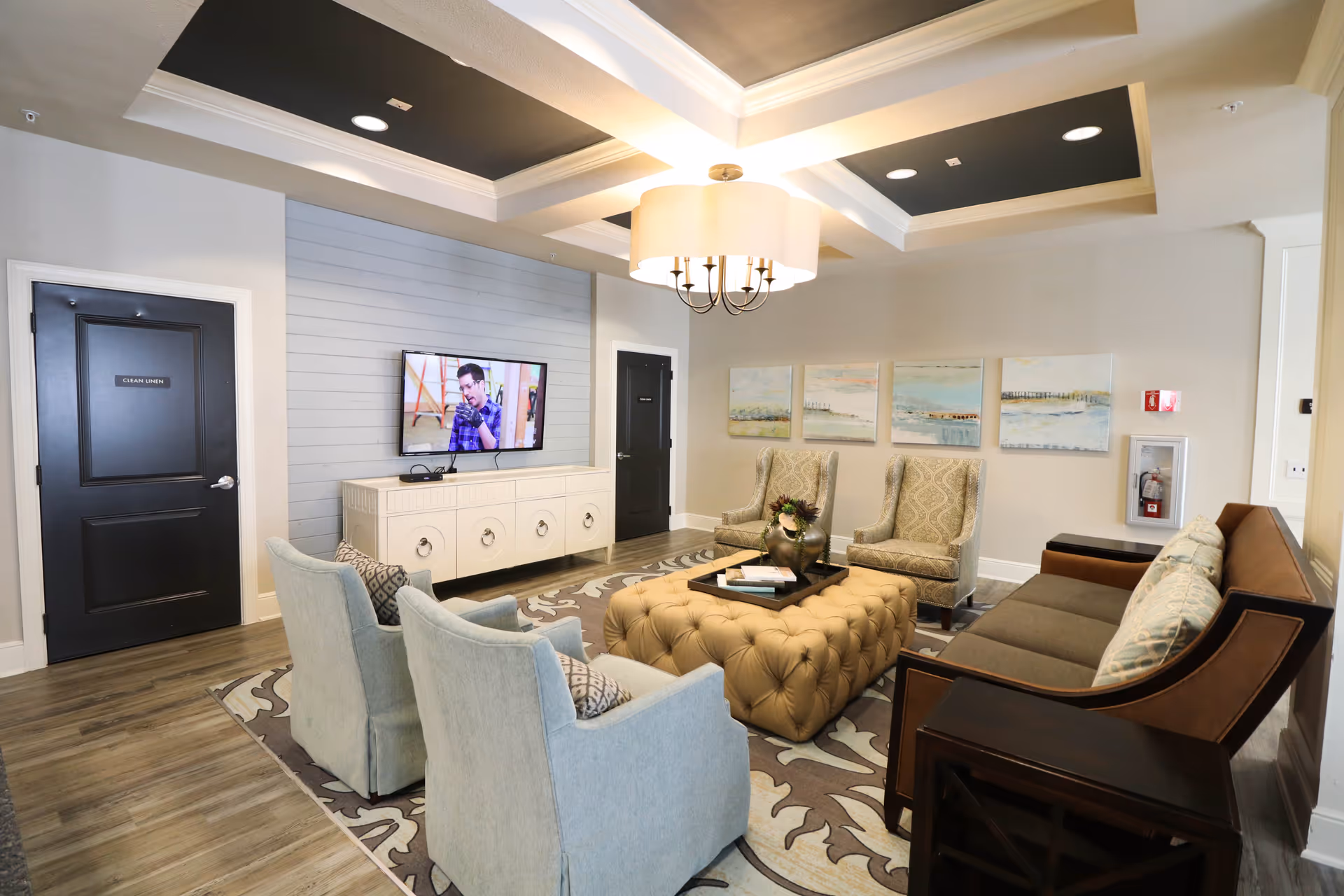 A cozy assisted living facility lounge with a tufted ottoman in the center, surrounded by two light blue armchairs, two patterned armchairs, and a brown sofa. A flat-screen TV is mounted on a light blue shiplap wall above a white cabinet. The room has wood flooring, a patterned area rug, and a ceiling light fixture with a large drum shade. There are four landscape paintings on the wall and two black doors labeled 'Clean Linen' and another door.
