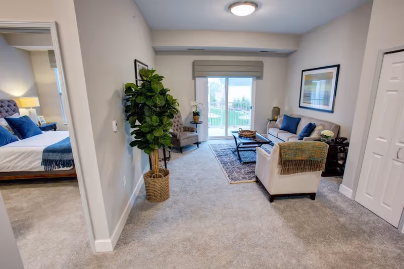 A cozy living room with a beige sofa, armchair with a throw blanket, coffee table on a patterned rug, and a large potted plant near the entrance. The room has a sliding glass door leading to an outdoor area. To the left, there is a view into a bedroom with a bed, nightstand, and lamp.