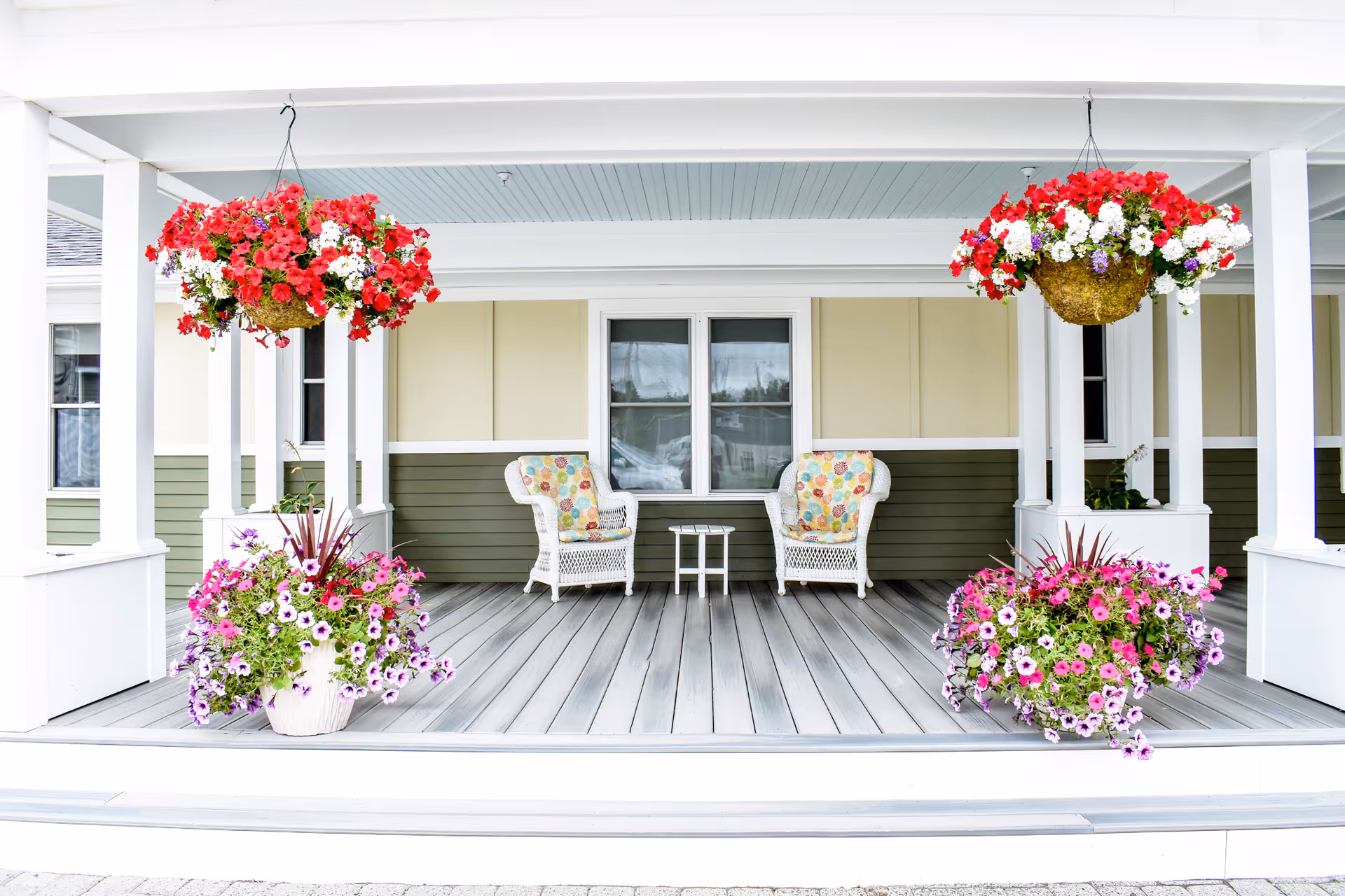 A covered front porch with two wicker chairs and a small table flanked by hanging baskets and potted flowering plants.