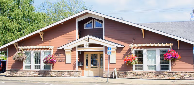 Exterior front view of a single-story building with brown siding and stone accents. The building has a peaked roof with small windows near the top and two large windows on either side of the entrance. Hanging flower baskets with vibrant pink and purple flowers are displayed on both sides of the entrance. A handicap parking sign is visible near the front door.