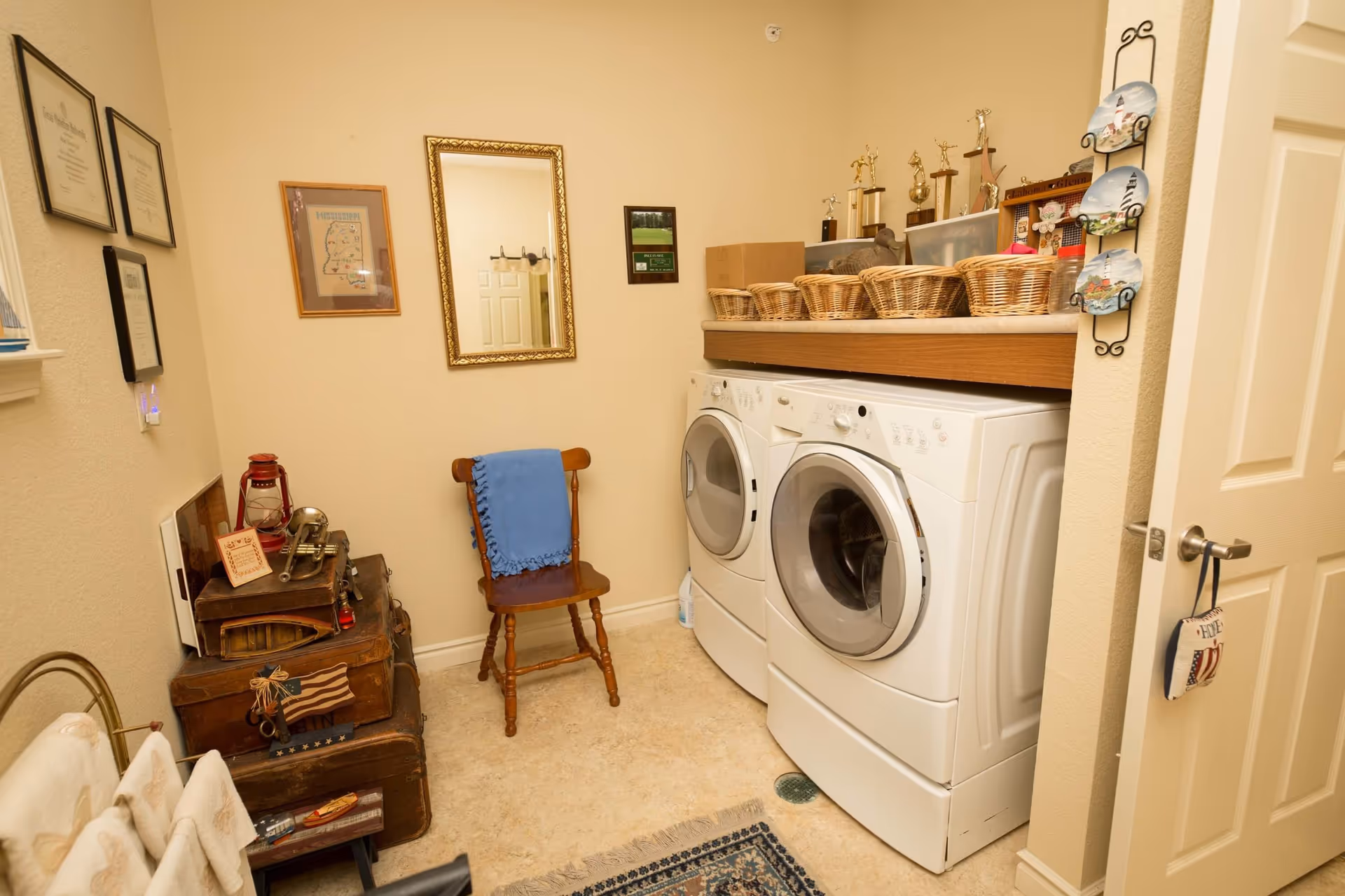 Laundry room with side-by-side front-loading washer and dryer, a shelf with wicker baskets, a wooden chair and decorative trunks.