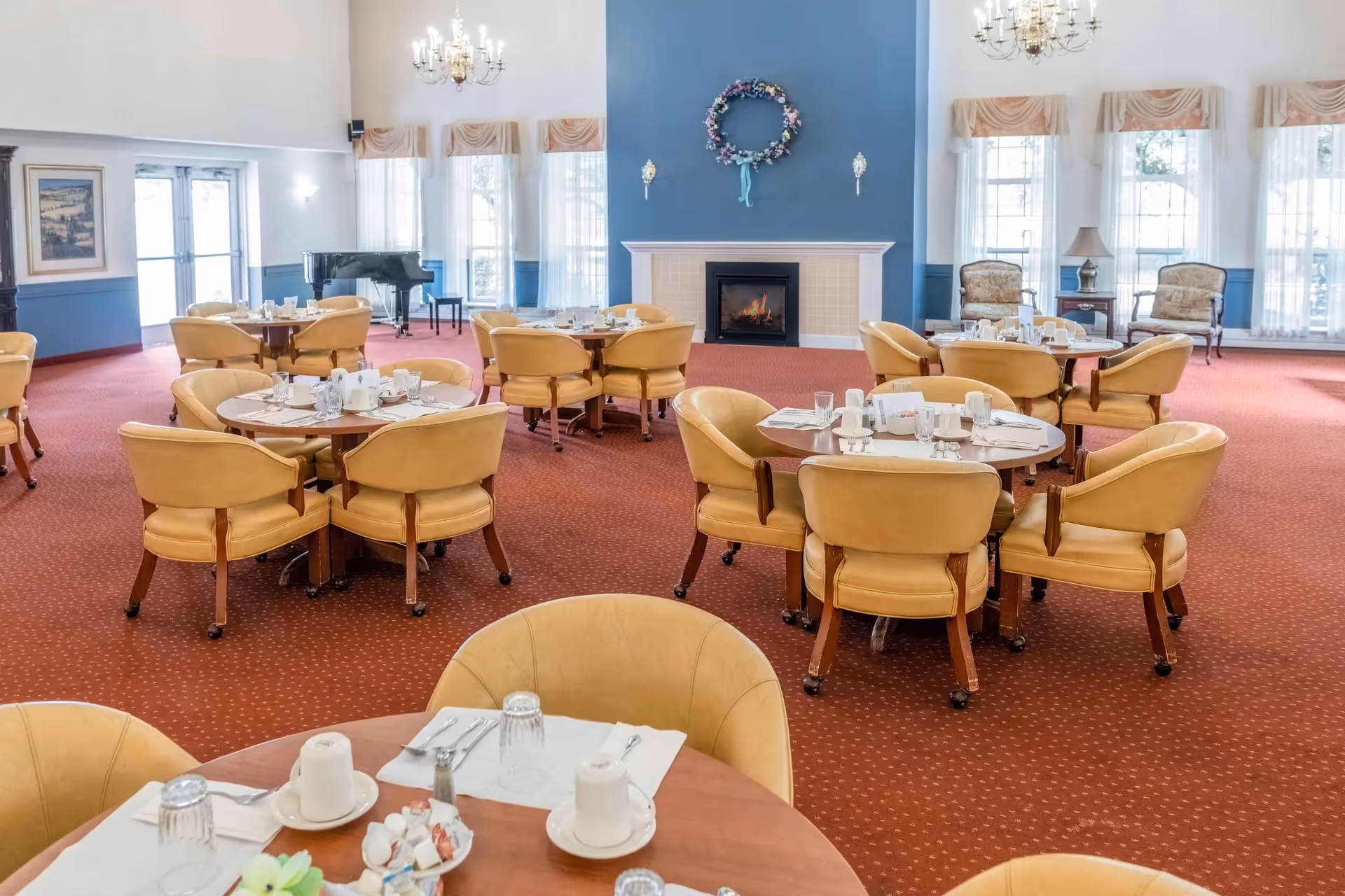A spacious dining room with round tables set for meals, surrounded by beige cushioned chairs on wheels. The room has a red carpet with a subtle pattern, large windows with sheer curtains and valances, a blue accent wall with a fireplace decorated with a floral wreath, chandeliers hanging from the ceiling, and a black grand piano in the corner.