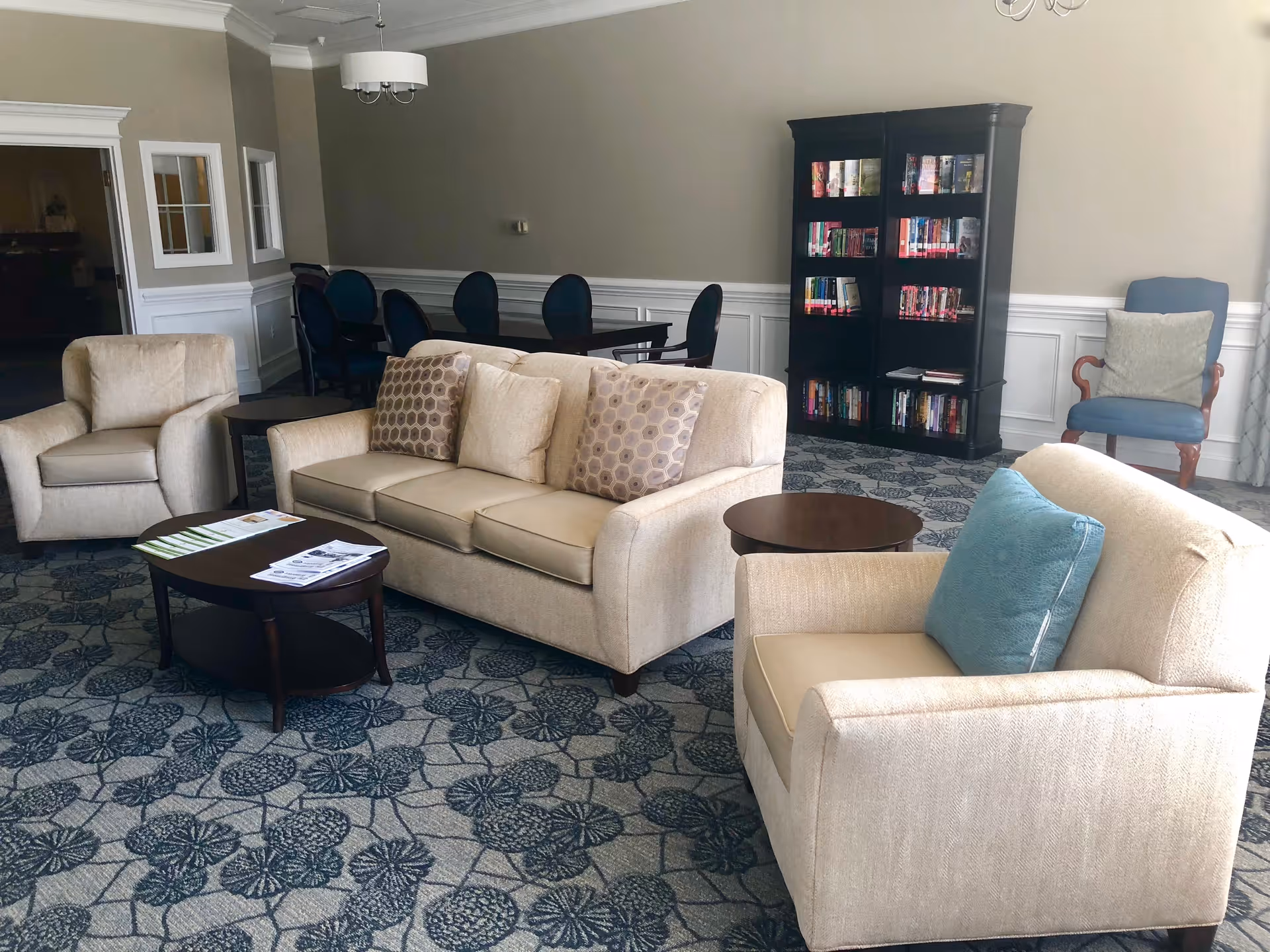 A cozy living room area in an assisted living facility with beige upholstered sofa and armchairs, a dark wooden coffee table with papers on it, a round side table, a patterned carpet, a black bookshelf filled with books, and a blue cushioned chair with a pillow near a window.