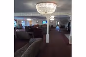 Interior view of a senior living facility lounge area with multiple upholstered chairs and sofas arranged around the room. A large chandelier hangs from the ceiling, and there are columns and decorative molding on the walls. A television is mounted on the far wall.