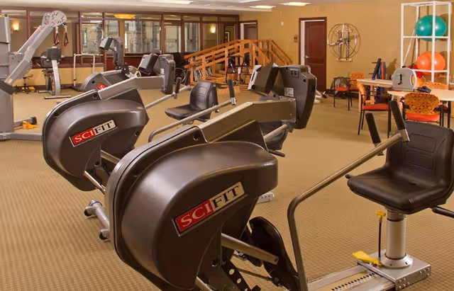 Interior fitness room with multiple SciFit exercise machines, therapy equipment, and seating in a senior living facility.