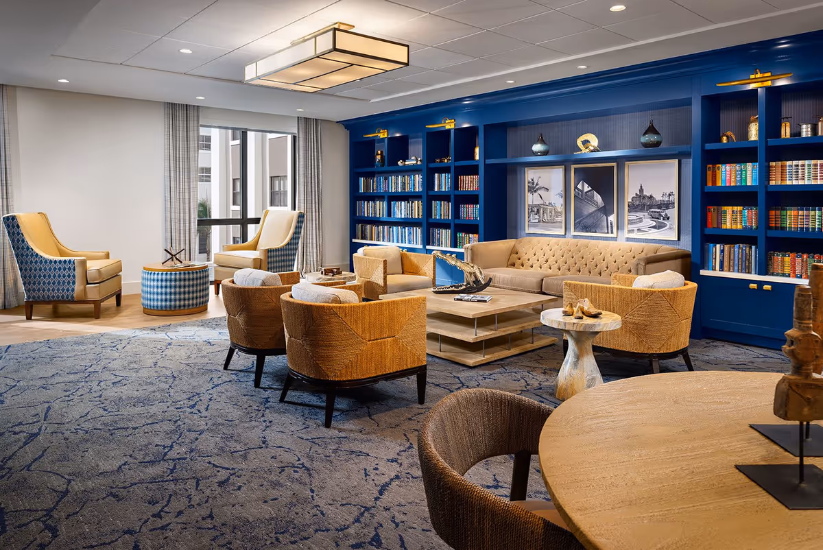 A bright communal lounge with blue built-in bookshelves, a tufted sofa, armchairs, coffee tables, and a patterned rug.