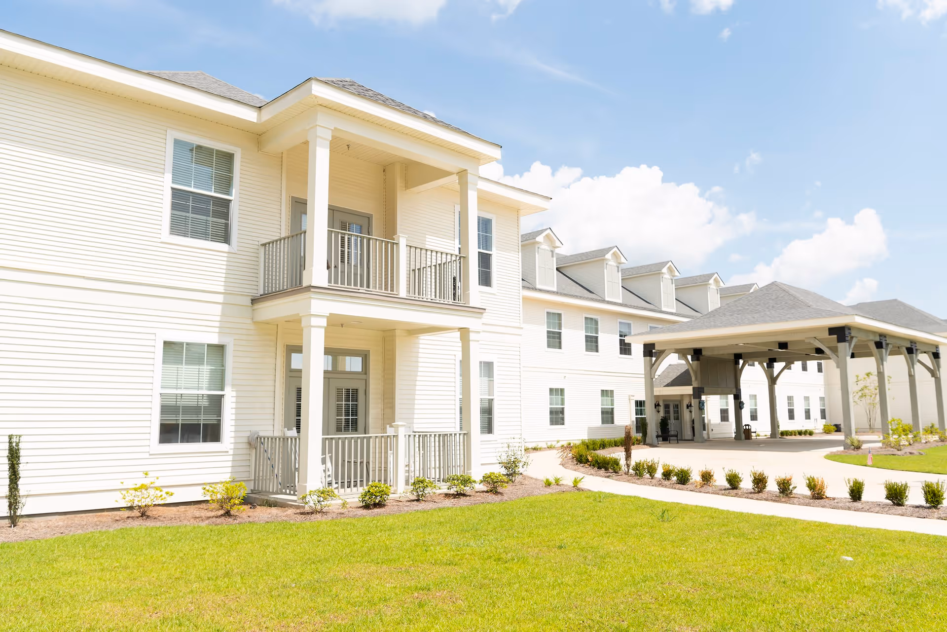 Exterior view of a senior living facility building with white siding, multiple windows, a covered entrance with pillars, and a well-maintained lawn and landscaping under a partly cloudy sky.