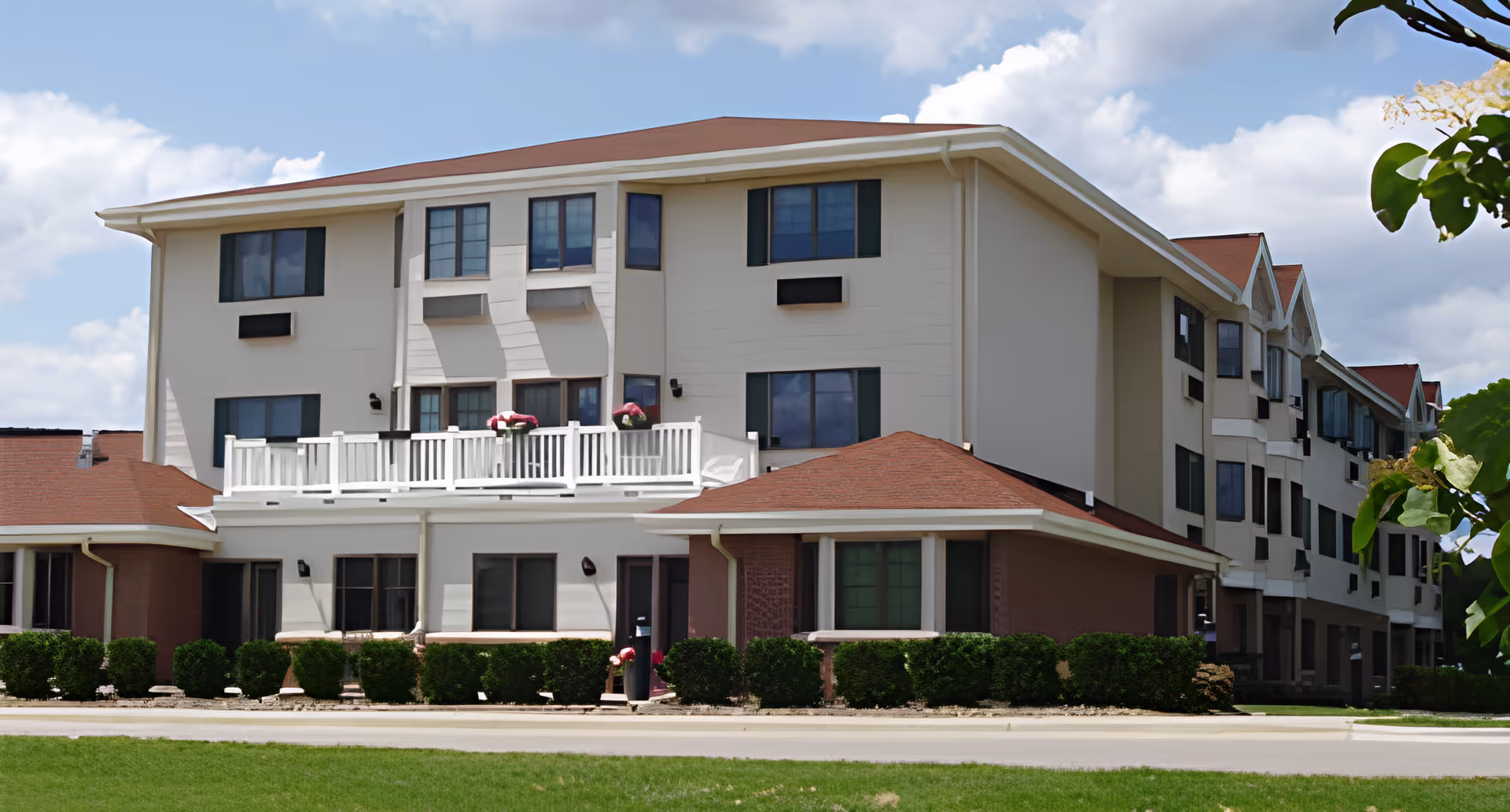 Exterior view of a multi-story retirement facility building with beige walls, red roofs, multiple windows, a white balcony with flower pots, and neatly trimmed bushes in front under a partly cloudy sky.