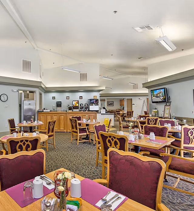 Spacious dining room with multiple set tables and upholstered chairs, a serving counter in the back, and a wall-mounted TV.
