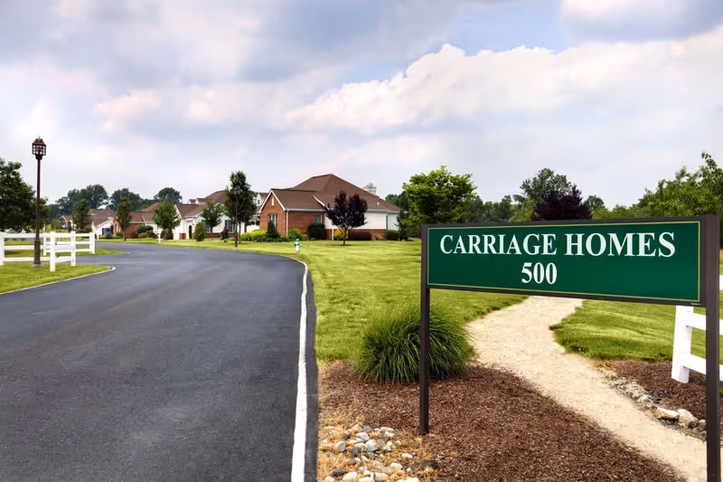 A paved road curves to the left with white fences and lamp posts along the side. On the right, there is a green sign that reads 'CARRIAGE HOMES 500' next to a gravel pathway leading to single-story brick homes surrounded by green lawns and trees under a partly cloudy sky.