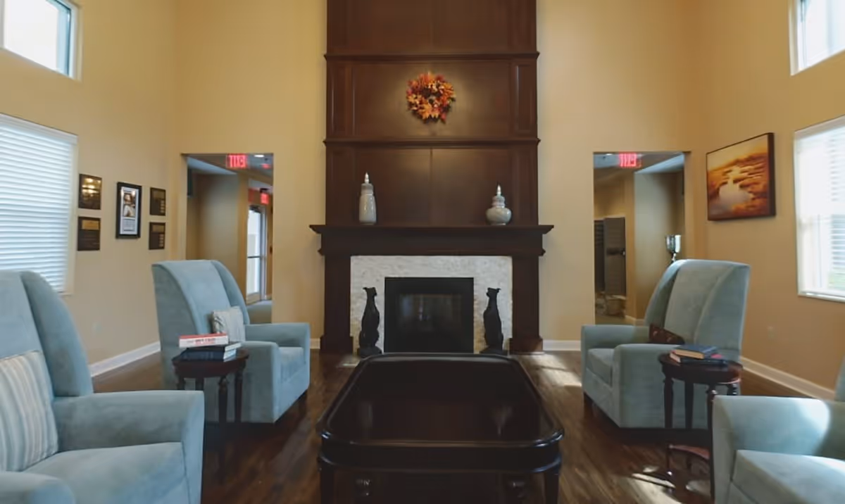 A cozy living room with four light blue armchairs arranged around a dark wooden coffee table. The room features a dark wooden fireplace with a white stone surround, decorated with two vases and a fall-themed wreath above it. There are windows on both sides letting in natural light, a painting on the right wall, and plaques on the left wall.