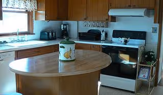 A kitchen with wooden cabinets, a white stove and oven, a microwave, a toaster, and a small island with a decorative container on top. There is a window with a floral valance above the sink.