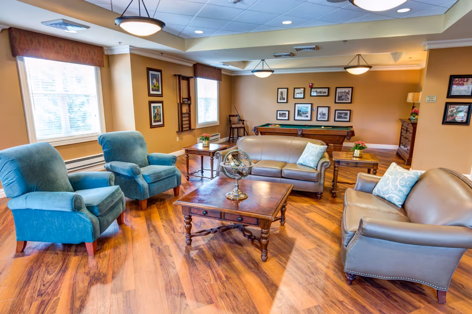 A cozy living room area in Symphony Manor featuring two blue armchairs, two brown leather sofas with decorative pillows, a wooden coffee table with a decorative globe, and side tables with small flower arrangements. The room has wooden flooring, beige walls adorned with framed pictures, and a pool table in the background. Large windows with valances allow natural light to fill the space.