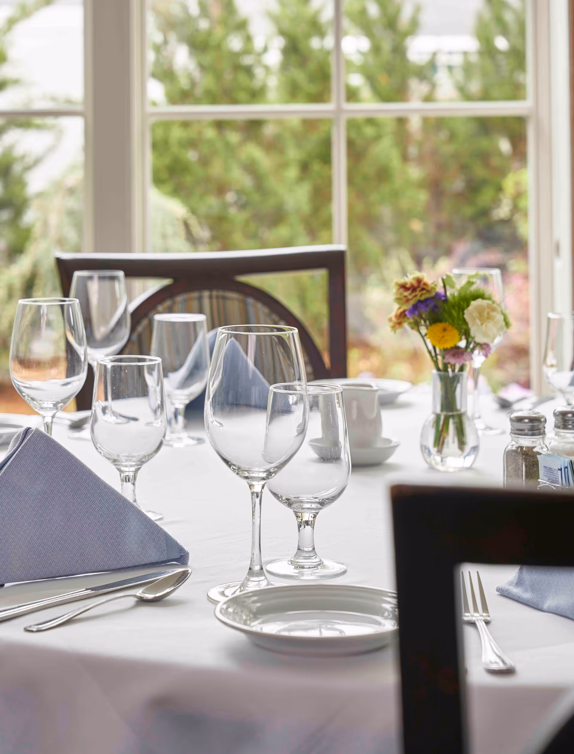 A neatly set dining table with wine glasses, folded napkins, plates and a small vase of flowers by a window.