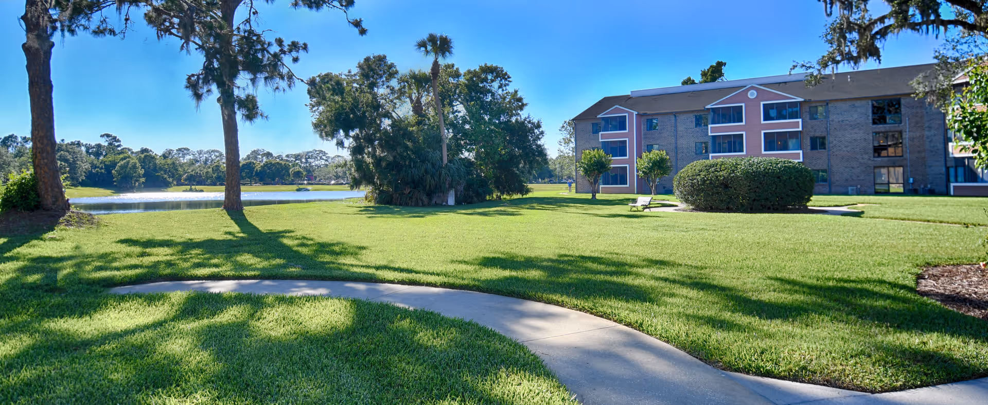 A sunny outdoor scene at CountrySide Lakes featuring a well-maintained grassy area with a curved concrete pathway, several trees including palm trees, a small pond in the background, and a three-story residential building with balconies on the right side.