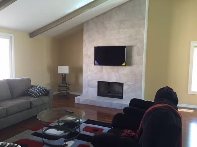 A living room with a sofa and recliner facing a tiled fireplace topped by a wall-mounted TV, with a glass coffee table on a patterned rug.