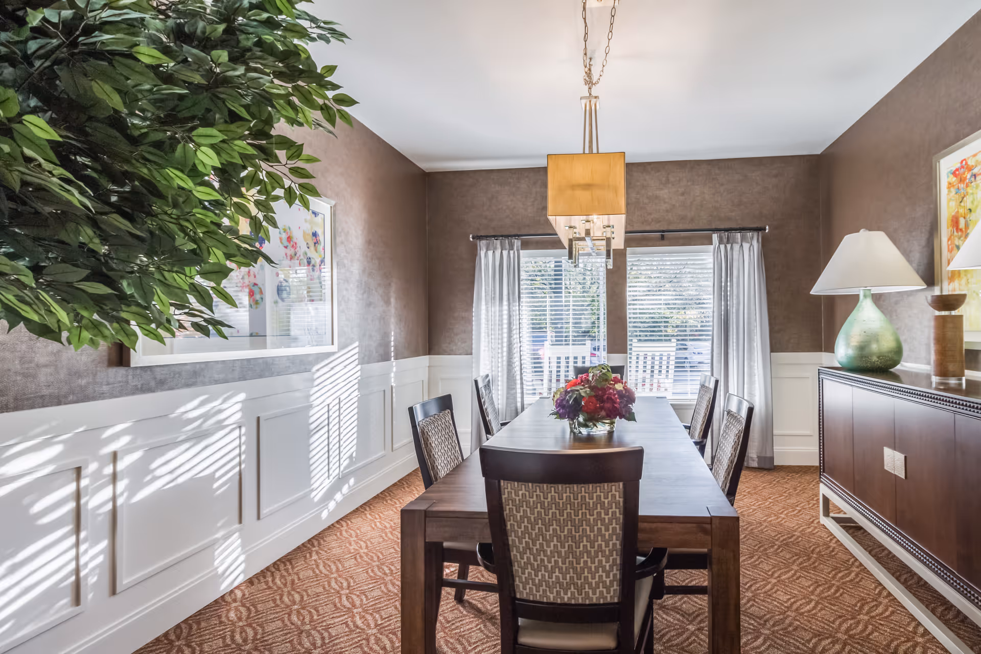 Sunlit dining room with a long wooden table and chairs, a chandelier overhead, sideboard with a lamp, and windows with curtains.