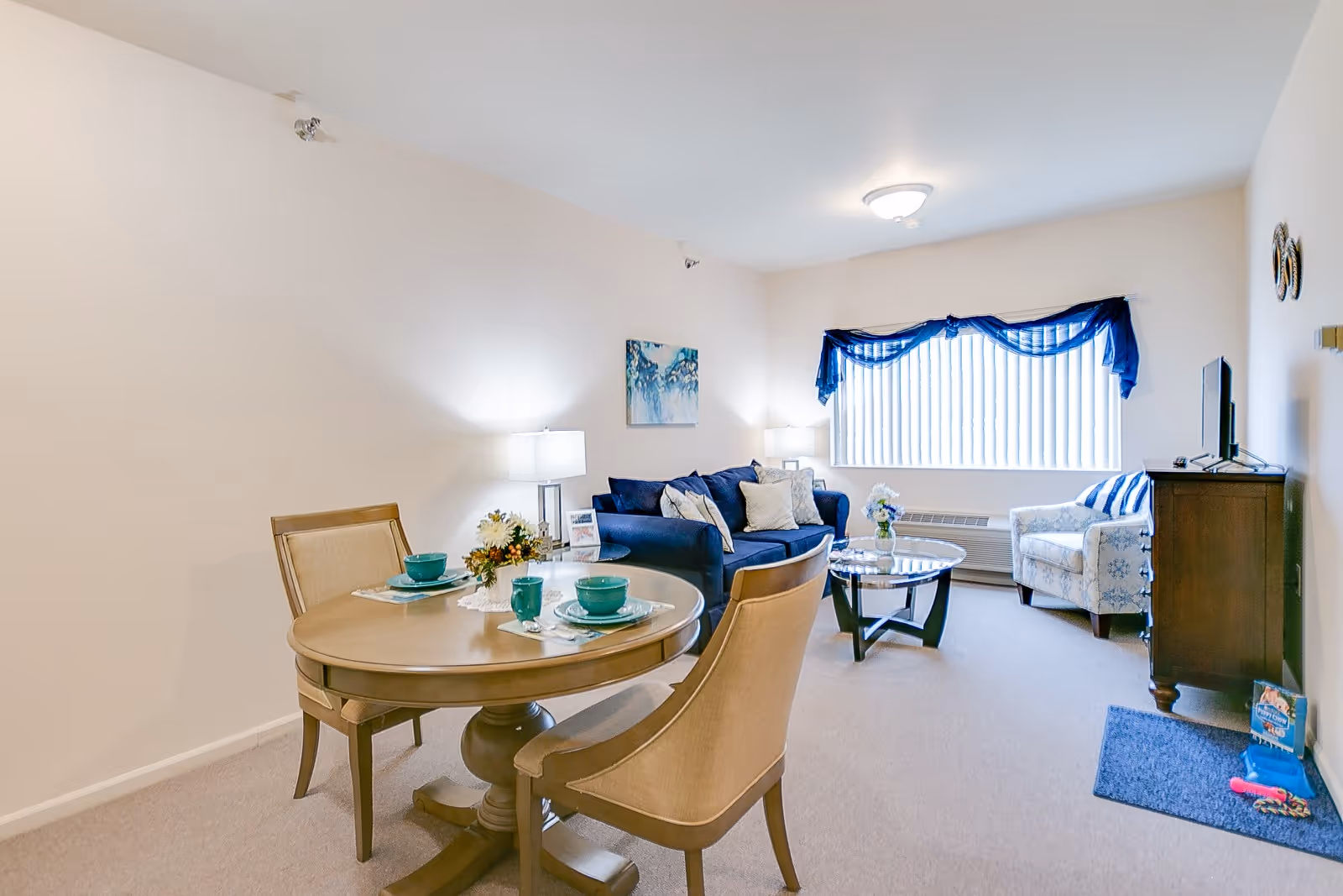 A bright living room and dining area in a senior living facility. The dining area has a round wooden table set with teal dishes and two chairs. The living room features a blue sofa with multiple cushions, a glass coffee table with a flower vase, a patterned armchair, a wooden TV stand with a flat-screen TV, and a large window with vertical blinds and a blue valance.