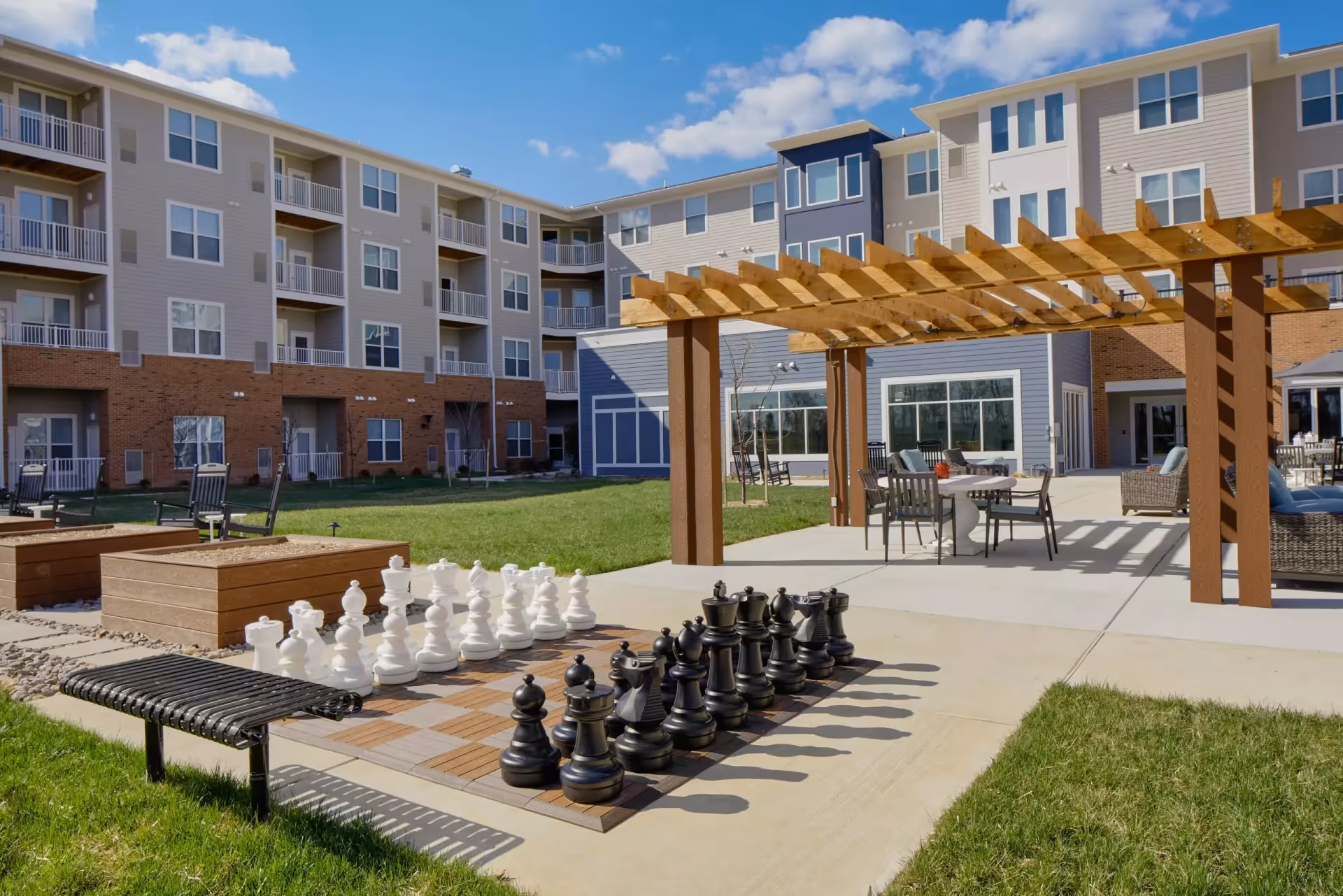 Outdoor courtyard area of a senior living facility with a large chessboard and oversized chess pieces, a black metal bench, wooden pergola with patio tables and chairs, and a multi-story building in the background under a partly cloudy sky.