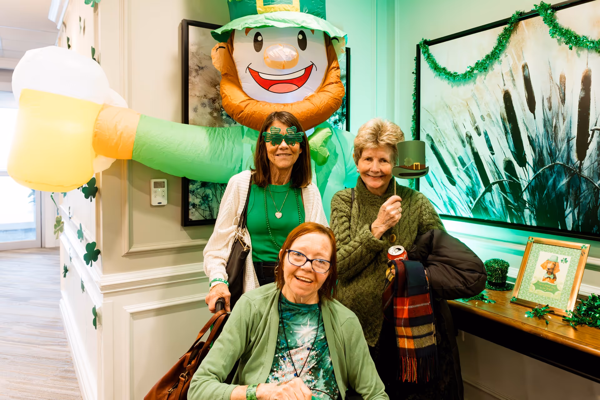 Three elderly women posing indoors in front of a large inflatable leprechaun decoration. The women are dressed in green and holding St. Patrick's Day themed props, including shamrock glasses and a small leprechaun hat on a stick. The room is decorated with green garlands and shamrock cutouts, creating a festive atmosphere.