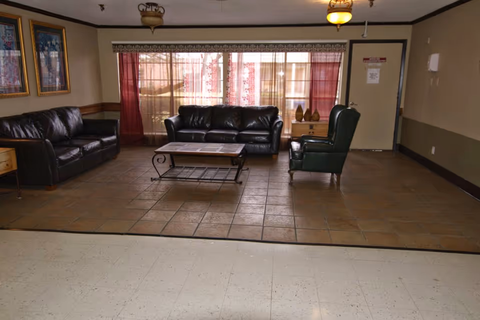 A common area with two black leather sofas, a green armchair, and a coffee table on a tiled floor. The background features large windows with sheer and red curtains, framed artwork on the wall, and a door to the right.
