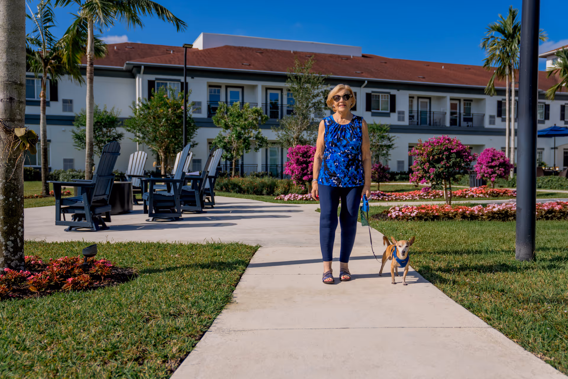 An elderly woman wearing sunglasses, a blue patterned sleeveless top, and navy pants is walking a small dog on a leash along a paved pathway in a landscaped garden area. The background shows a two-story senior living facility building with balconies, surrounded by green grass, palm trees, and colorful flowering bushes under a clear blue sky.