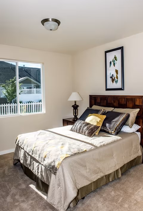 A bright bedroom with a large window showing an outdoor view of a white picket fence and neighboring houses. The room features a neatly made bed with multiple decorative pillows and a patterned bedspread. A wooden headboard, a bedside table with a lamp, and a framed artwork on the wall complete the cozy setting.