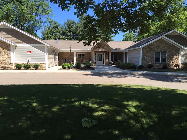 Front exterior view of a single-story building with beige siding and stone accents, surrounded by green trees and a well-maintained lawn under a clear blue sky.