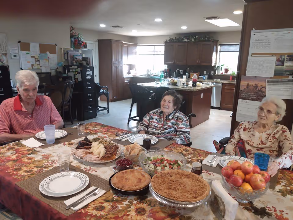 Three elderly people sitting around a dining table set with plates, glasses, and various dishes including pies, salad, rolls, and cranberry sauce in a kitchen and dining area.