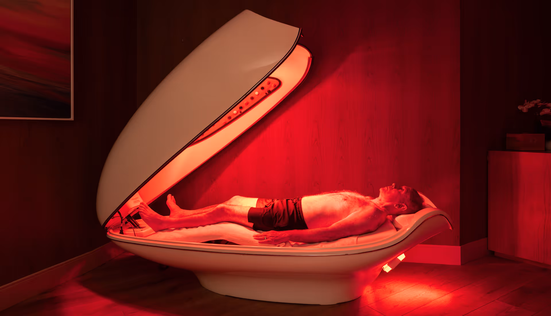 A man lying inside a futuristic pod-like relaxation or therapy capsule emitting red light, in a dimly lit room with wooden walls and floor.