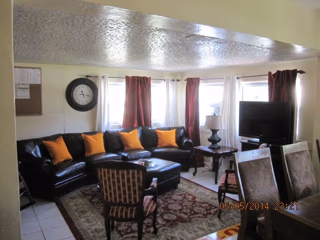 A cozy living room with a black leather sectional sofa adorned with orange and yellow throw pillows, a patterned armchair, a dark wooden coffee table, and a side table with a lamp. The room has three windows with red and white curtains, a large wall clock, and a flat-screen TV on a stand. A dining table with chairs is partially visible in the foreground.