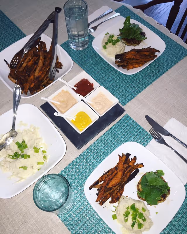 A dining table set with two plates of food, each containing mashed potatoes garnished with green onions, a serving of sweet potato fries, and a lettuce-topped patty. There are two glasses of water, a bowl of additional sweet potato fries with tongs, and four small square dishes containing ketchup, mustard, and two types of creamy sauces. The table has a beige tablecloth and turquoise woven placemats.