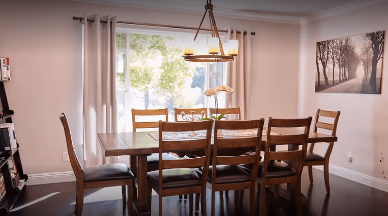 Wooden dining table with six chairs beneath a chandelier in a bright room with a large window and curtains.