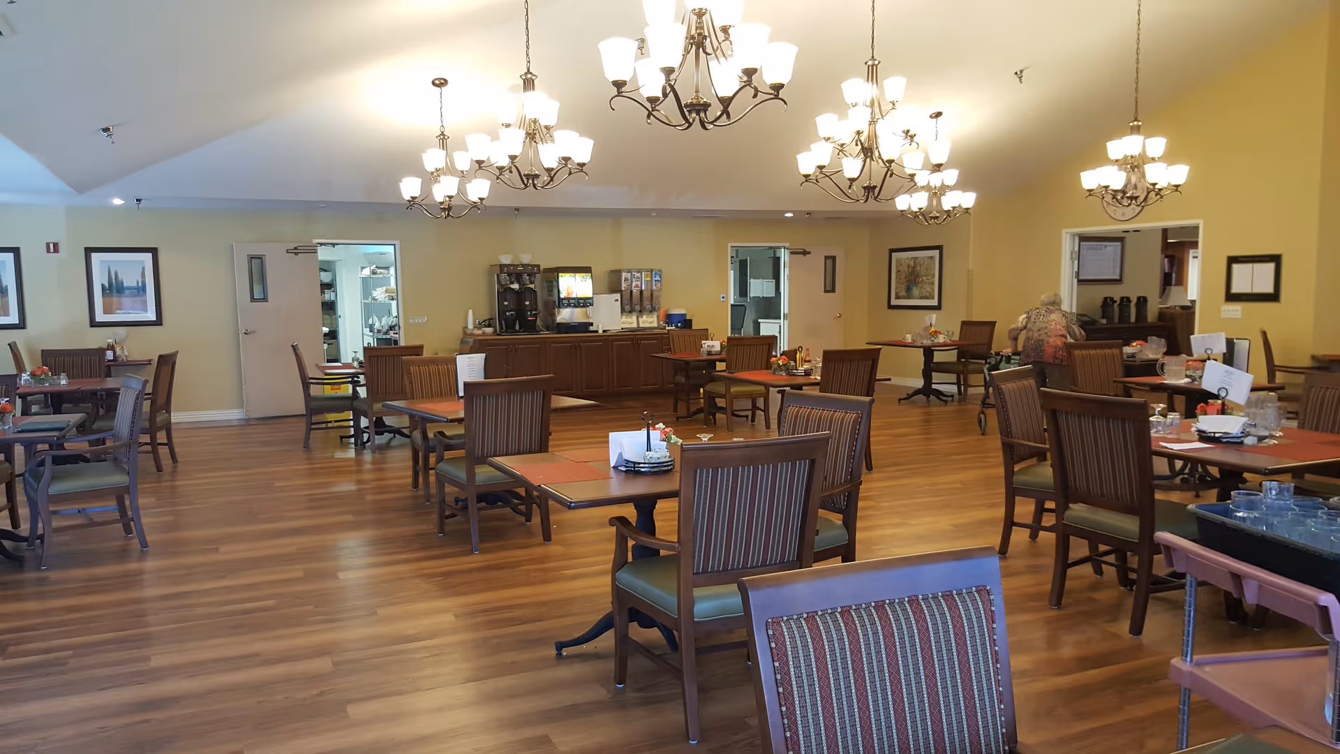 A spacious dining room with wooden floors and multiple tables and chairs arranged neatly. The room has soft yellow walls, several chandeliers hanging from the ceiling, and framed artwork on the walls. There is a counter area at the back with beverage dispensers and other items. A person is seated at one of the tables on the right side of the room.