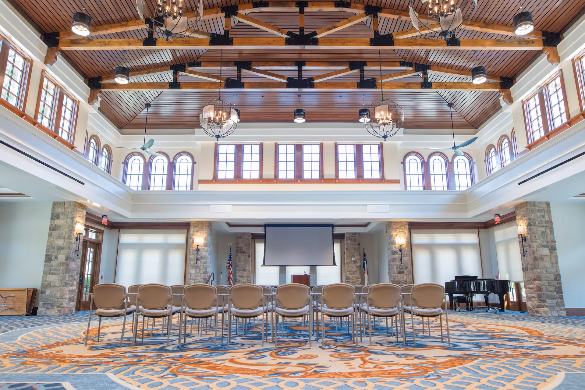 A spacious meeting or event room with a high wooden ceiling featuring exposed beams and chandeliers. The room has large windows with arched tops allowing natural light to fill the space. There are rows of beige chairs arranged facing a projector screen and podium at the front. Stone pillars and a grand piano are visible on the right side of the room. The carpet has an intricate blue and orange pattern.