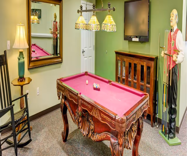 A game room with a small ornate pool table featuring a pink felt surface, a set of billiard balls arranged on the table, and a single cue ball. The room has green walls, a wooden rocking chair with a side table and lamp, a large mirror reflecting part of the room, a wall-mounted flat screen TV above a wooden cabinet, and a life-sized statue of a man holding pool cues standing in the corner.