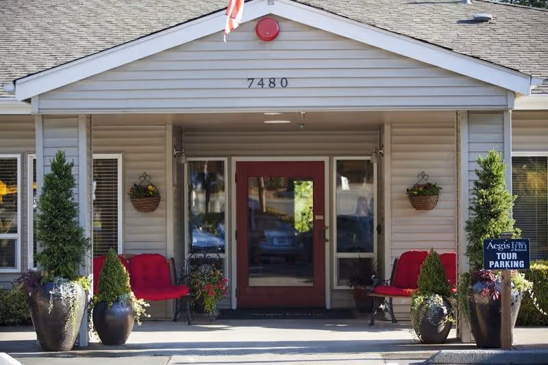 Entrance of a senior living facility with the address number 7480 displayed above the door. The entrance has a covered porch with two red cushioned benches on either side, large potted plants, and hanging flower baskets. A sign on the right side reads 'Aegis Living TOUR PARKING'.