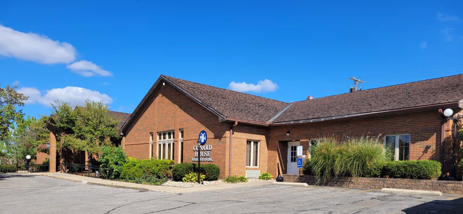 Exterior view of Conard House Assisted Living facility, a single-story brick building with a sloped roof, surrounded by shrubs and greenery under a clear blue sky.