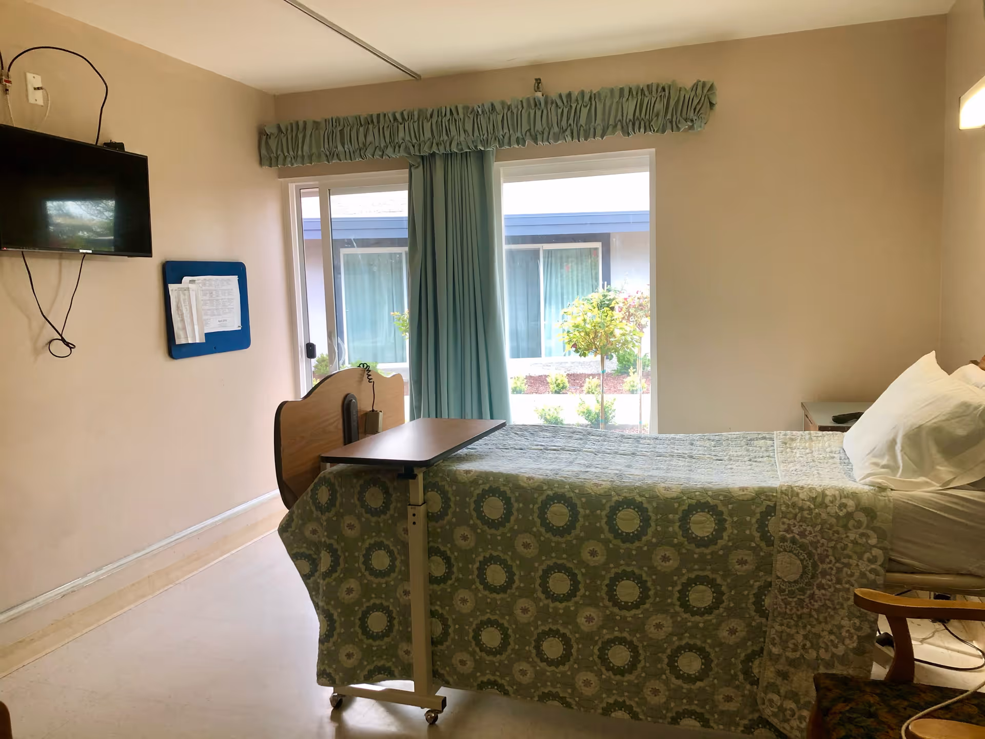 A simple patient room with a single bed covered with a green patterned bedspread, a small overbed table, a wall-mounted TV, a bulletin board with papers, a window with blue curtains, and a chair partially visible in the corner.