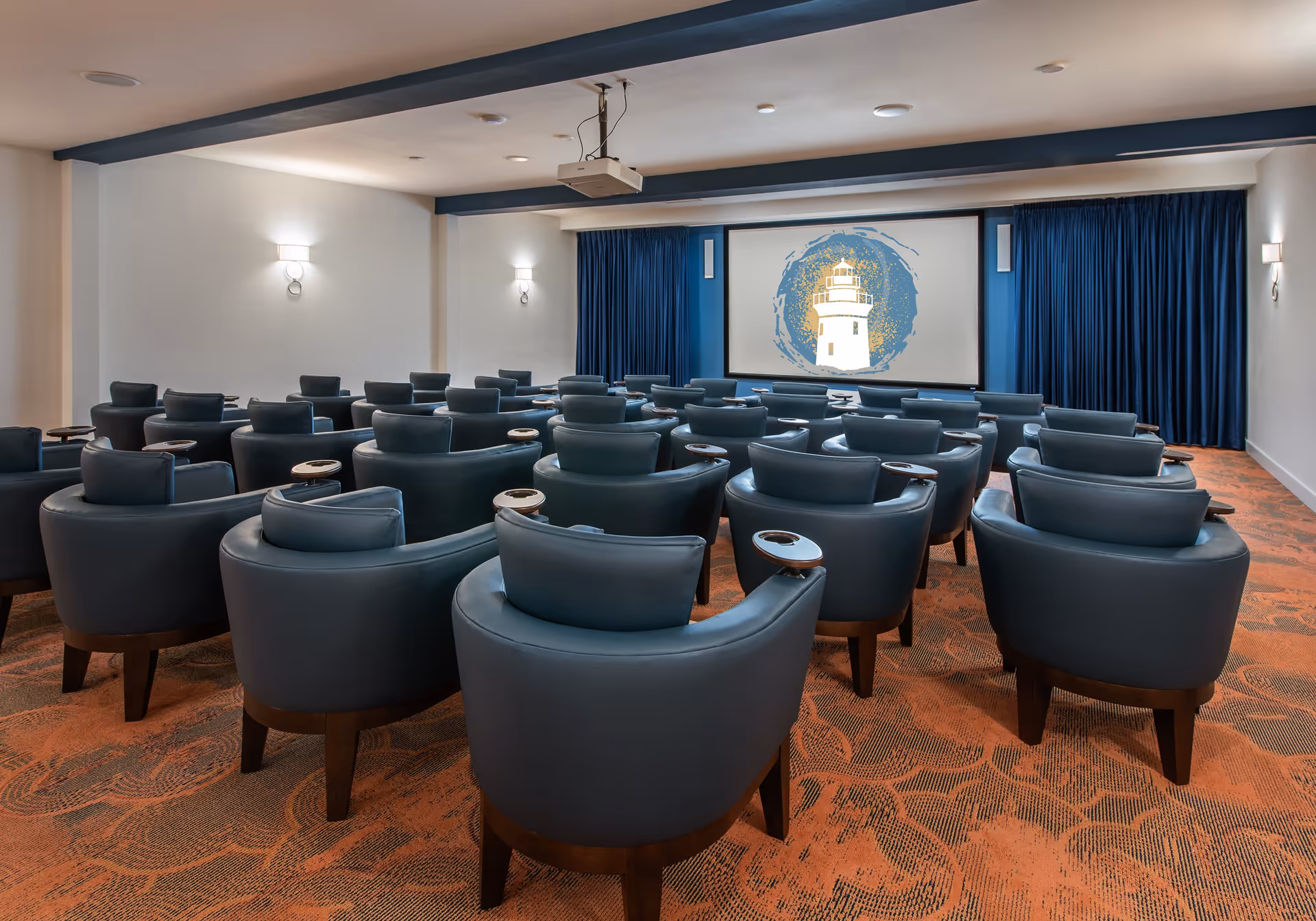 A small theater room with multiple rows of blue cushioned chairs with wooden legs and armrests, facing a large screen displaying an image of a lighthouse with a blue and gold circular background. The room has white walls with blue trim and blue curtains on either side of the screen, and orange patterned carpet.