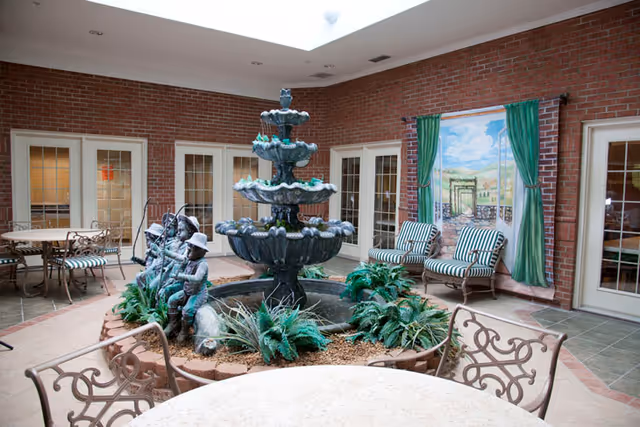 Indoor seating area with a large three-tiered fountain in the center surrounded by plants and a statue of children fishing. There are metal chairs and tables around the fountain, and cushioned chairs against a brick wall with a mural of a garden scene with green curtains painted on the wall.