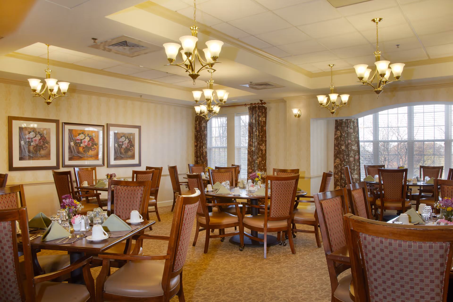 A well-lit dining room with multiple wooden tables and chairs arranged neatly. Each table is set with green folded napkins, white cups, glasses, and silverware. The room features beige patterned wallpaper, three framed floral paintings on one wall, and large windows with floral curtains allowing natural light to enter. Several elegant chandeliers hang from the ceiling providing warm lighting.