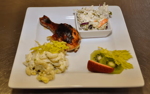 A white square plate with a grilled chicken drumstick, a serving of macaroni salad garnished with celery leaves, a small bowl of coleslaw, and slices of kiwi and strawberry garnished with celery leaves.