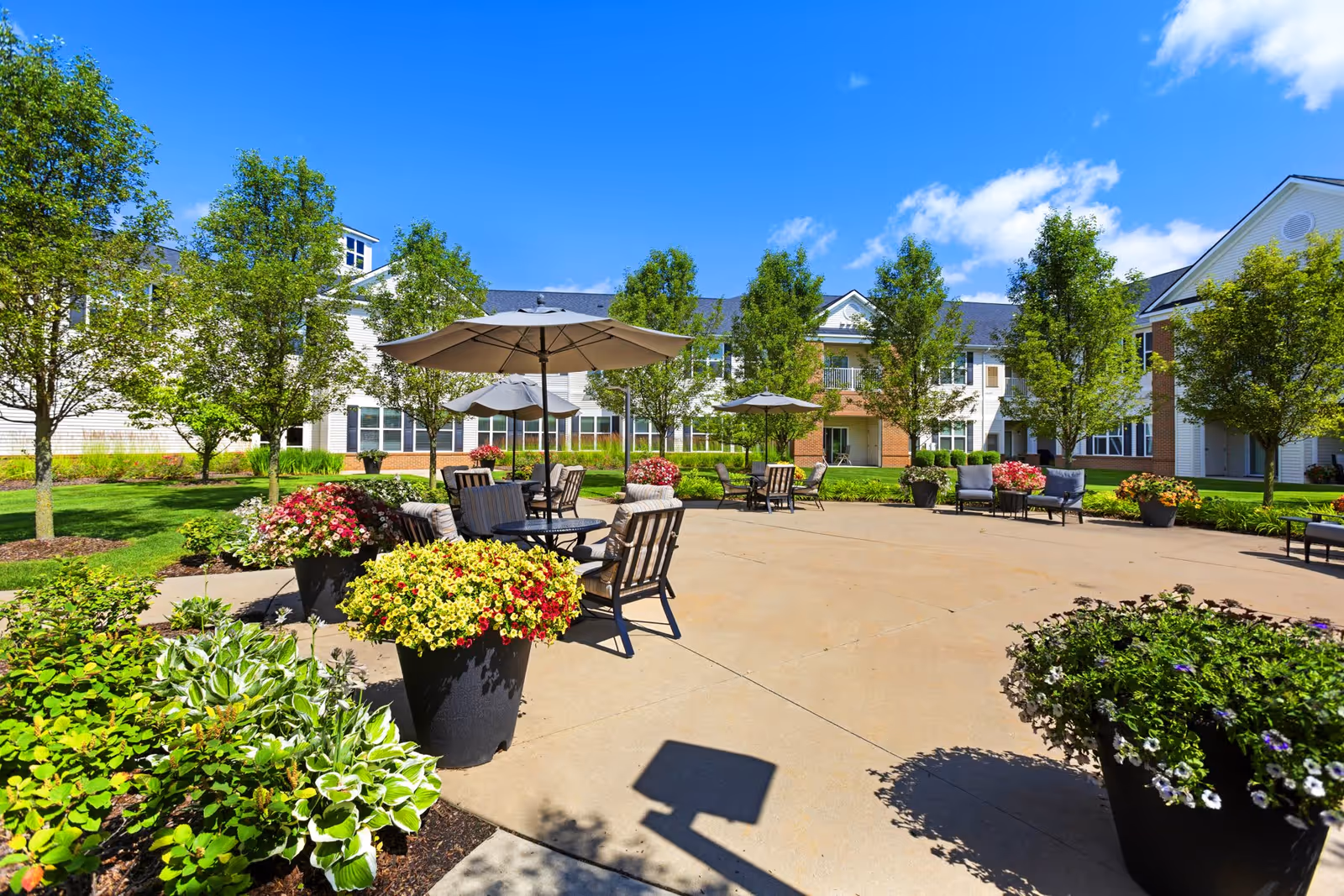 Outdoor patio area at Olivia's Assisted Living with several tables and chairs under large umbrellas, surrounded by potted flowers and greenery, with a building and trees in the background under a clear blue sky.