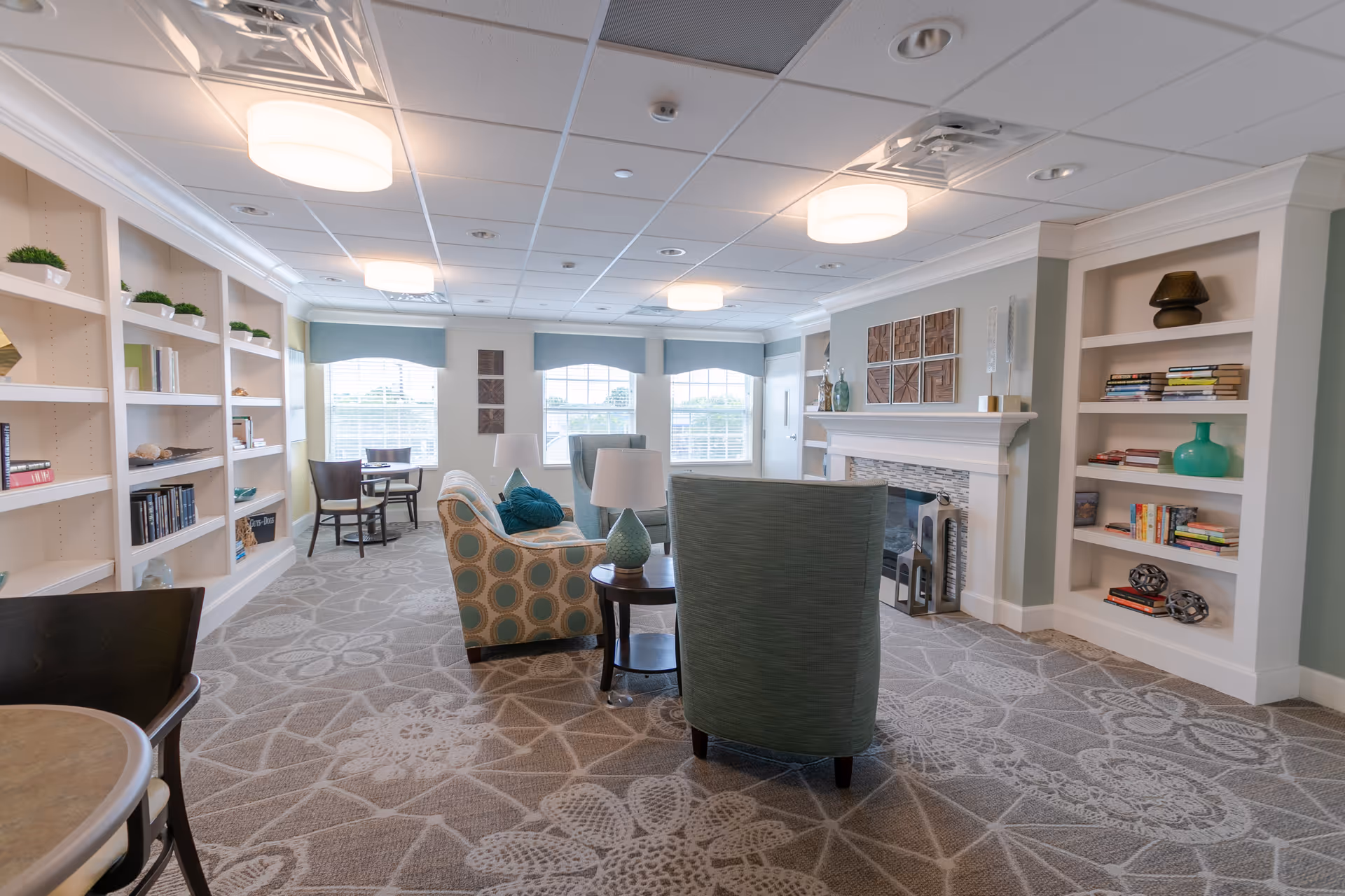 A bright and spacious senior living common area with patterned carpet, built-in bookshelves filled with books and decorative items, comfortable armchairs and a sofa arranged around a small round table with lamps, large windows with blue valances letting in natural light, and a white fireplace with decorative art above it.