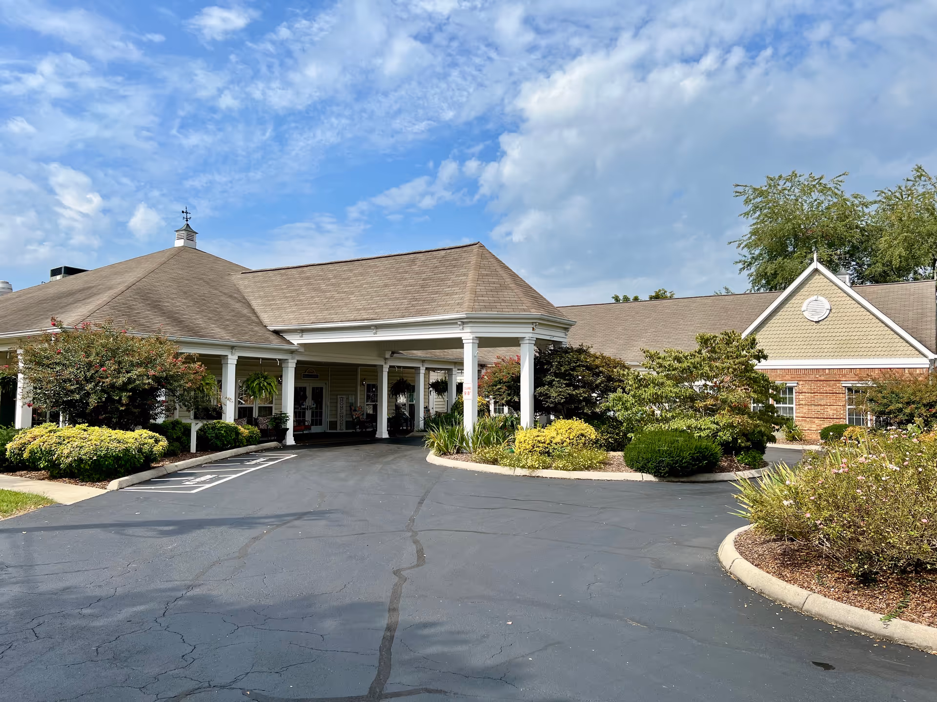 Entrance and driveway of a single-story building with a covered porte-cochere and landscaped beds under a partly cloudy sky.