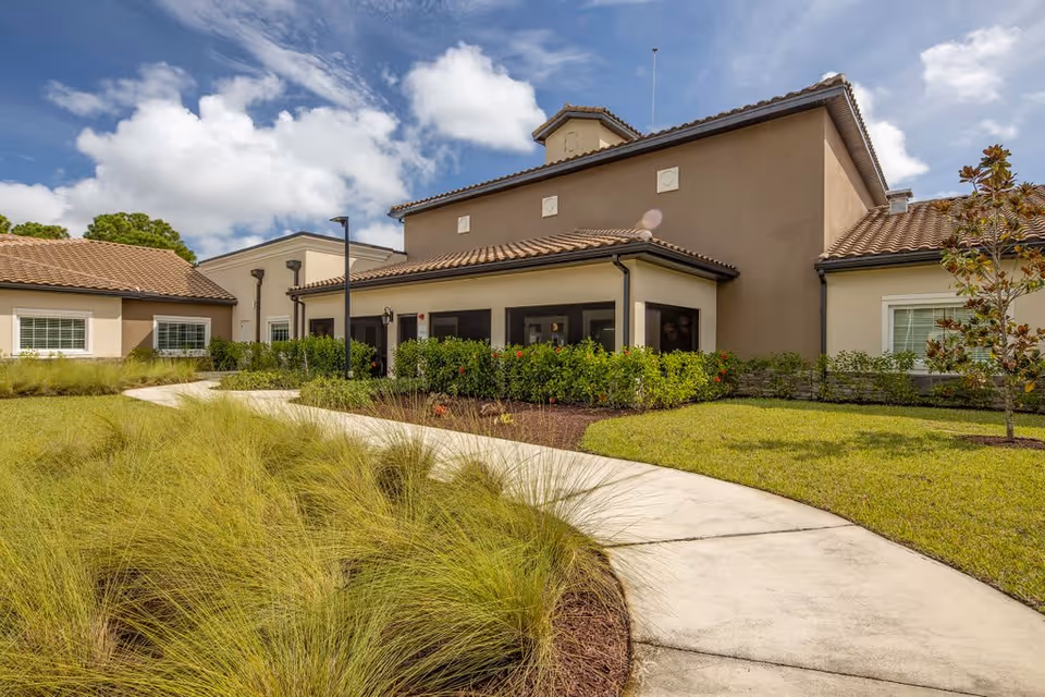 Exterior view of Discovery Village At Stuart showing a beige building with a tiled roof, surrounded by green grass, bushes, and a curved concrete walkway under a partly cloudy sky.
