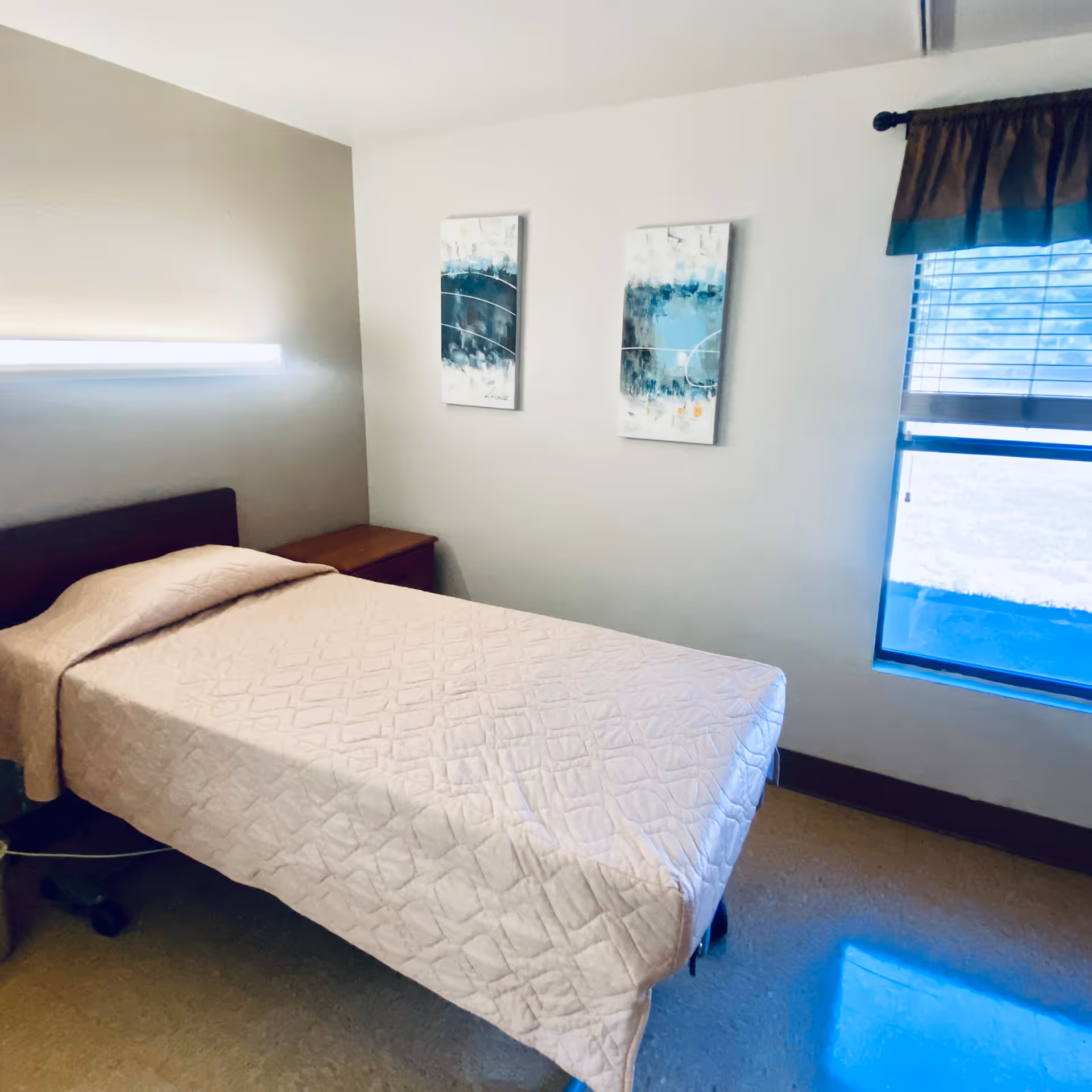 Small bedroom with a single bed, nightstand, two abstract wall paintings, and a window with blinds.